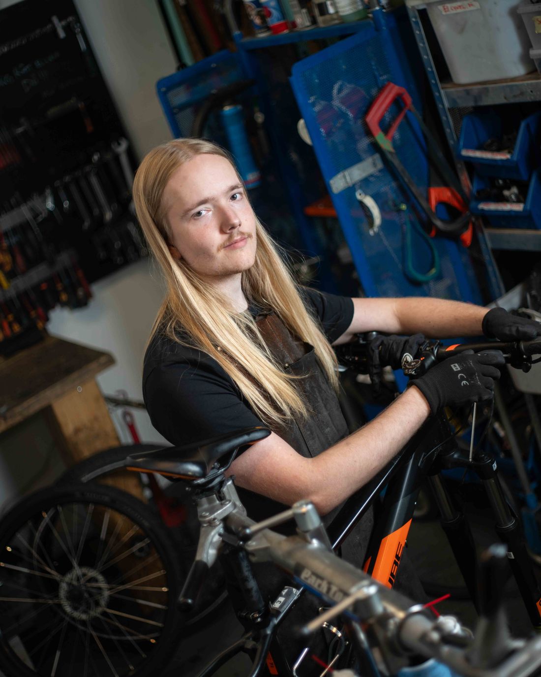 Mekaniker med långt hår och arbetshandskar går igenom en svart cykel vid verktygshyllorna